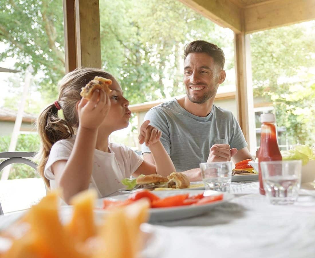 A man and a young girl sit at an outdoor table, smiling and eating hamburgers together. Sunlight filters through trees in the background, and there are plates, glasses, and a bottle of ketchup on the table.