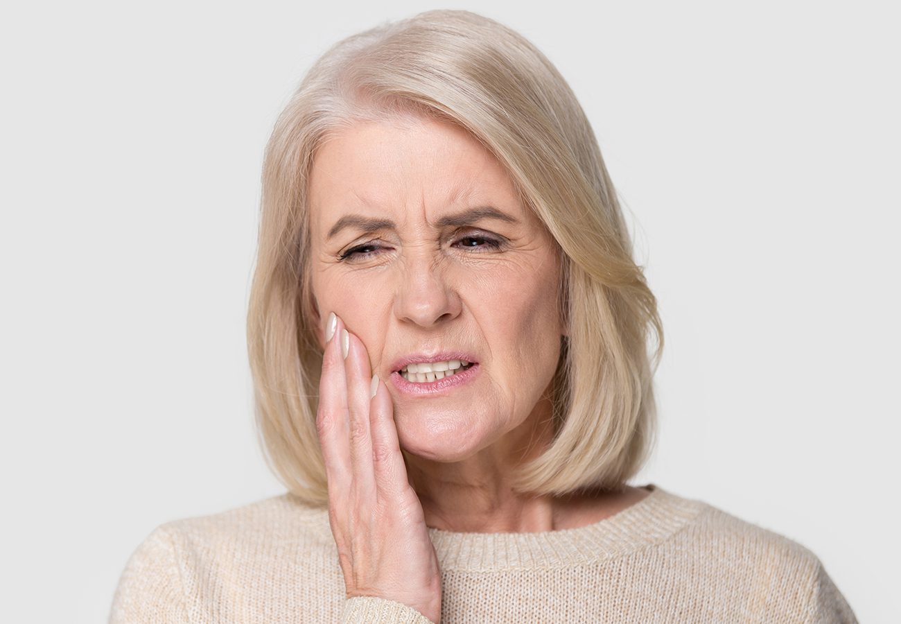 Older woman with short blond hair holds her hand to her cheek and winces in pain, possibly experiencing a toothache or jaw discomfort, against a plain light background.
