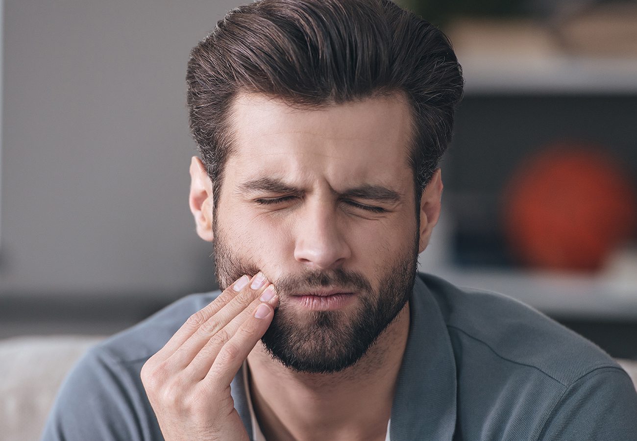 A man with a beard is touching his cheek and wincing in pain, possibly experiencing a toothache or jaw discomfort.