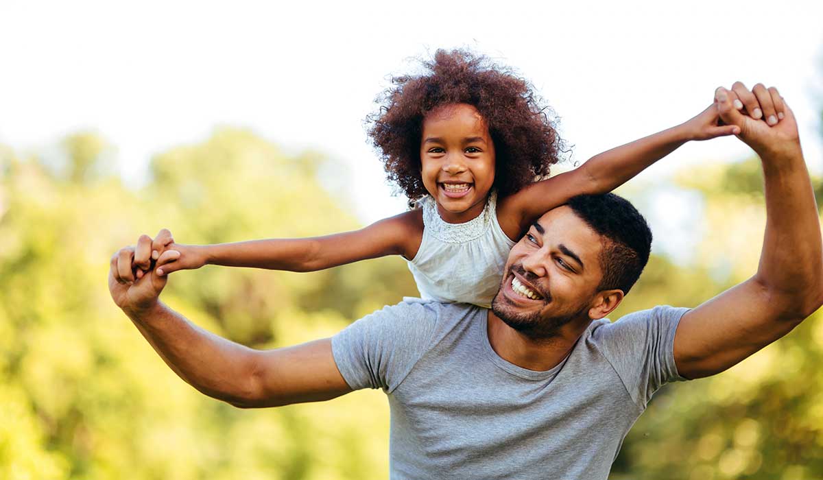 A smiling man in a gray shirt carries a cheerful young girl on his shoulders. They hold hands and pose outdoors against a blurred, green natural background.
