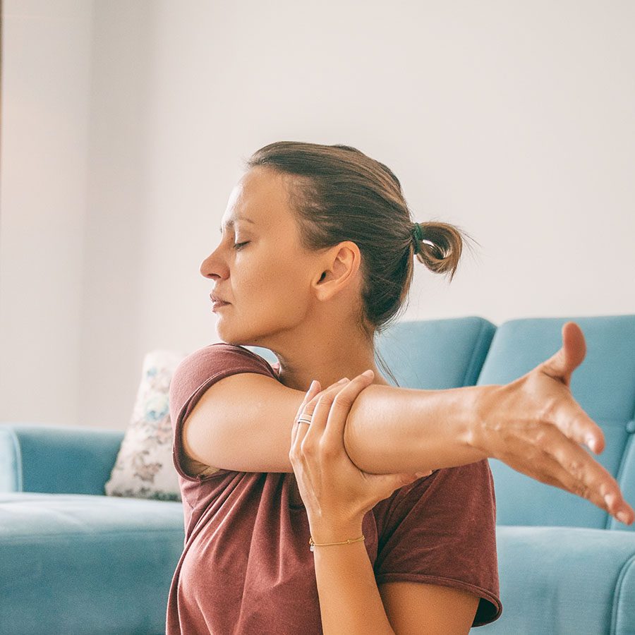 A woman with her hair in a ponytail stretches her right arm across her chest while sitting indoors, eyes closed, appearing relaxed. A blue couch is visible in the background.