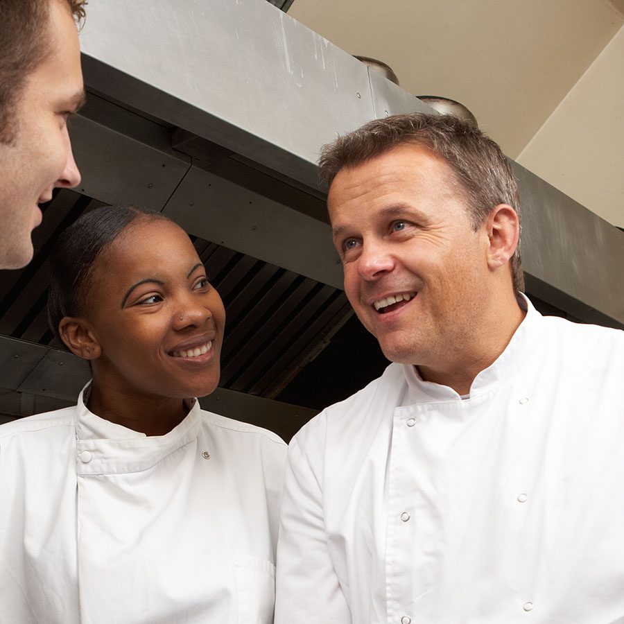 Two chefs wearing white uniforms smile and talk together in a professional kitchen, with industrial vents visible in the background.
