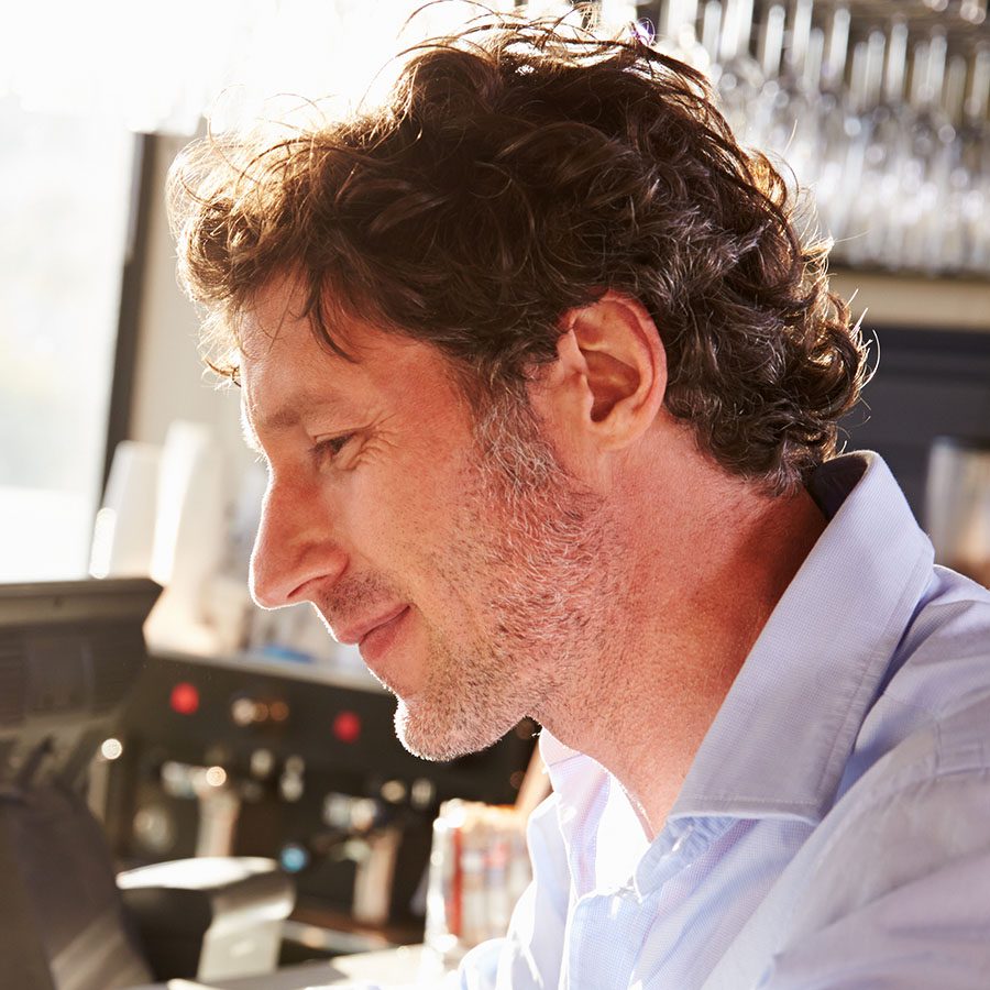 A man with curly brown hair and stubble, wearing a light blue shirt, is sitting indoors at a counter, looking down and smiling slightly in a warmly lit setting.