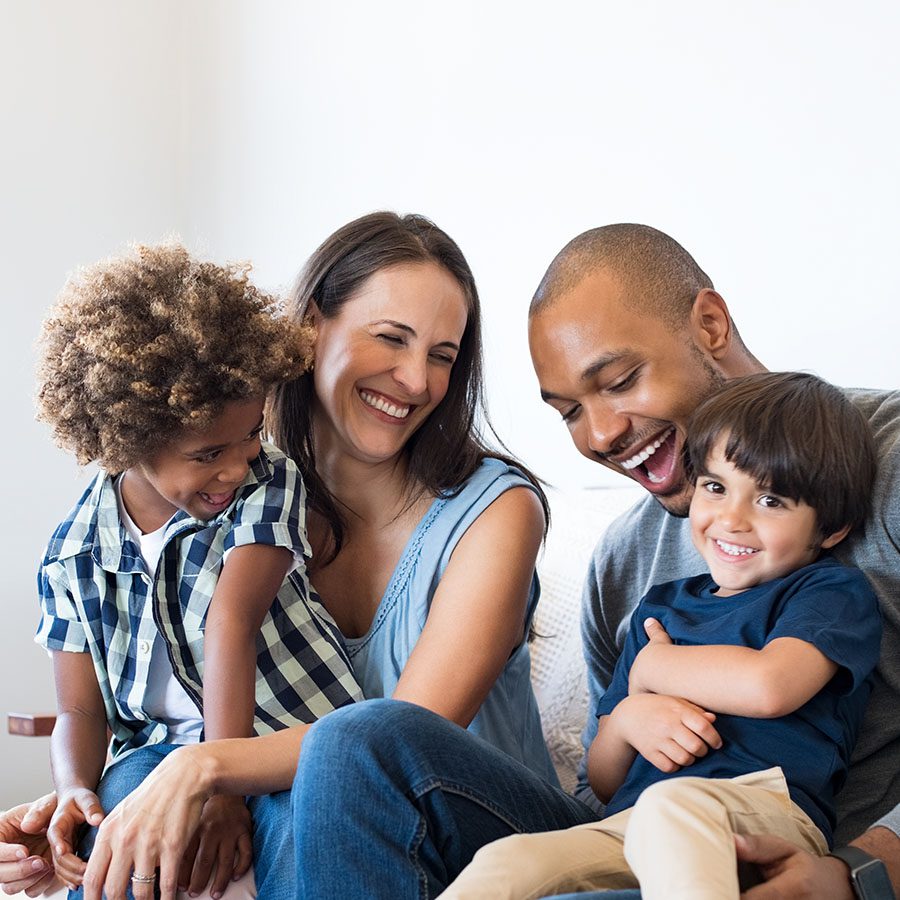 A smiling family of four, including two adults and two young children, sit closely together on a couch, laughing and enjoying each other’s company in a bright, cozy room.