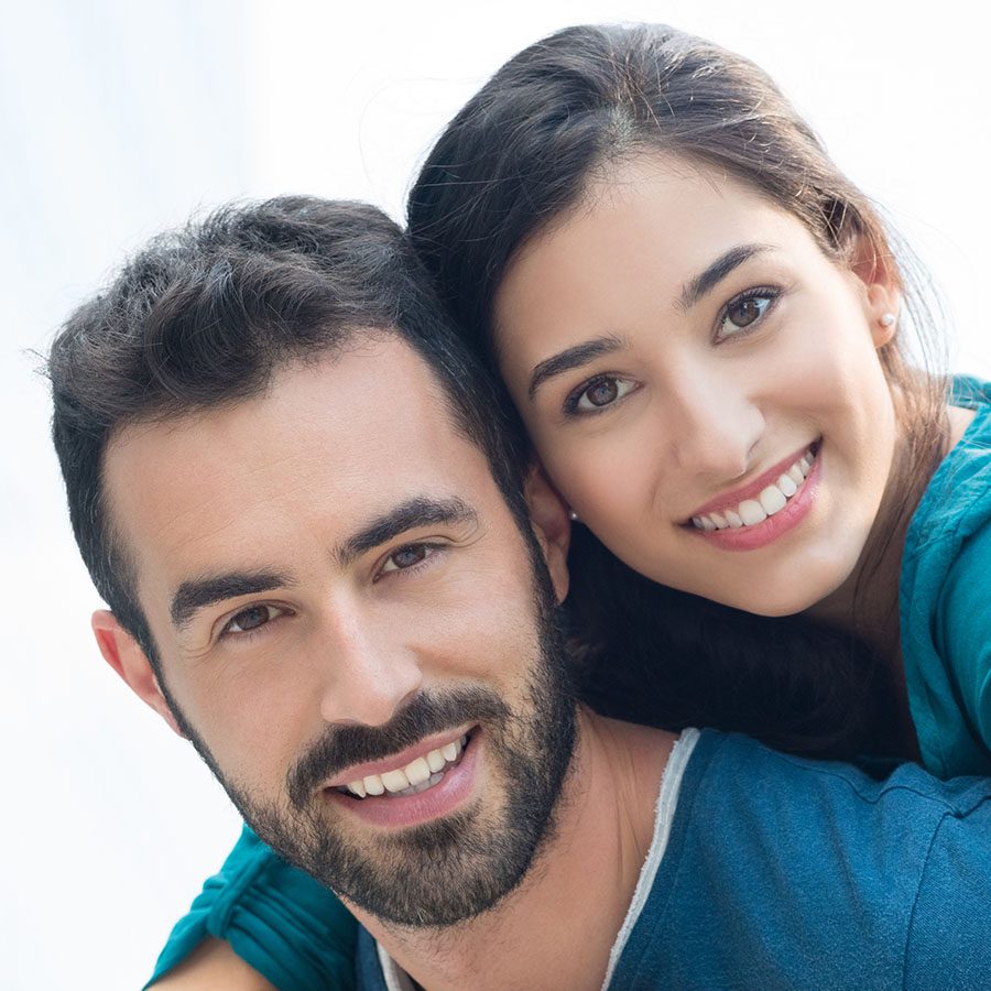 A smiling man with short dark hair and a beard is closely embraced by a smiling woman with long dark hair. Both are wearing teal tops and looking directly at the camera, showing their teeth. The background is bright and soft.