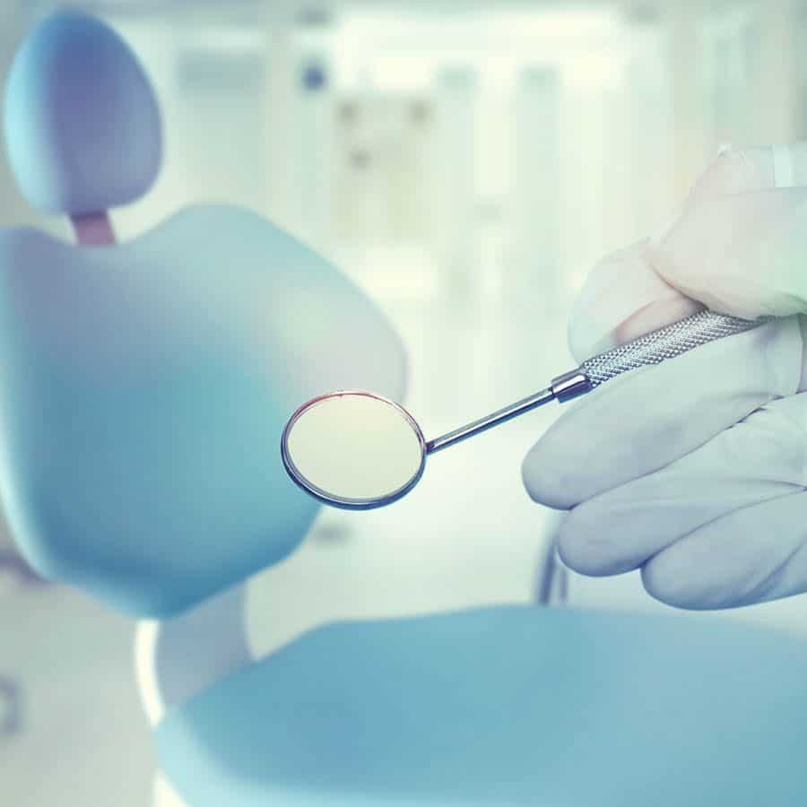A gloved hand holds a dental mirror in front of a blurred dentist chair and clinical background.