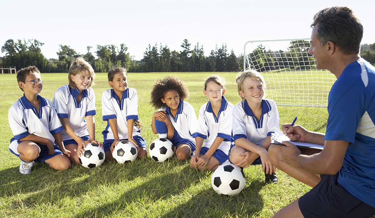 A group of six children in soccer uniforms sit on grass with soccer balls, smiling and listening to a coach who is kneeling and talking to them near a goalpost on a sunny field.
