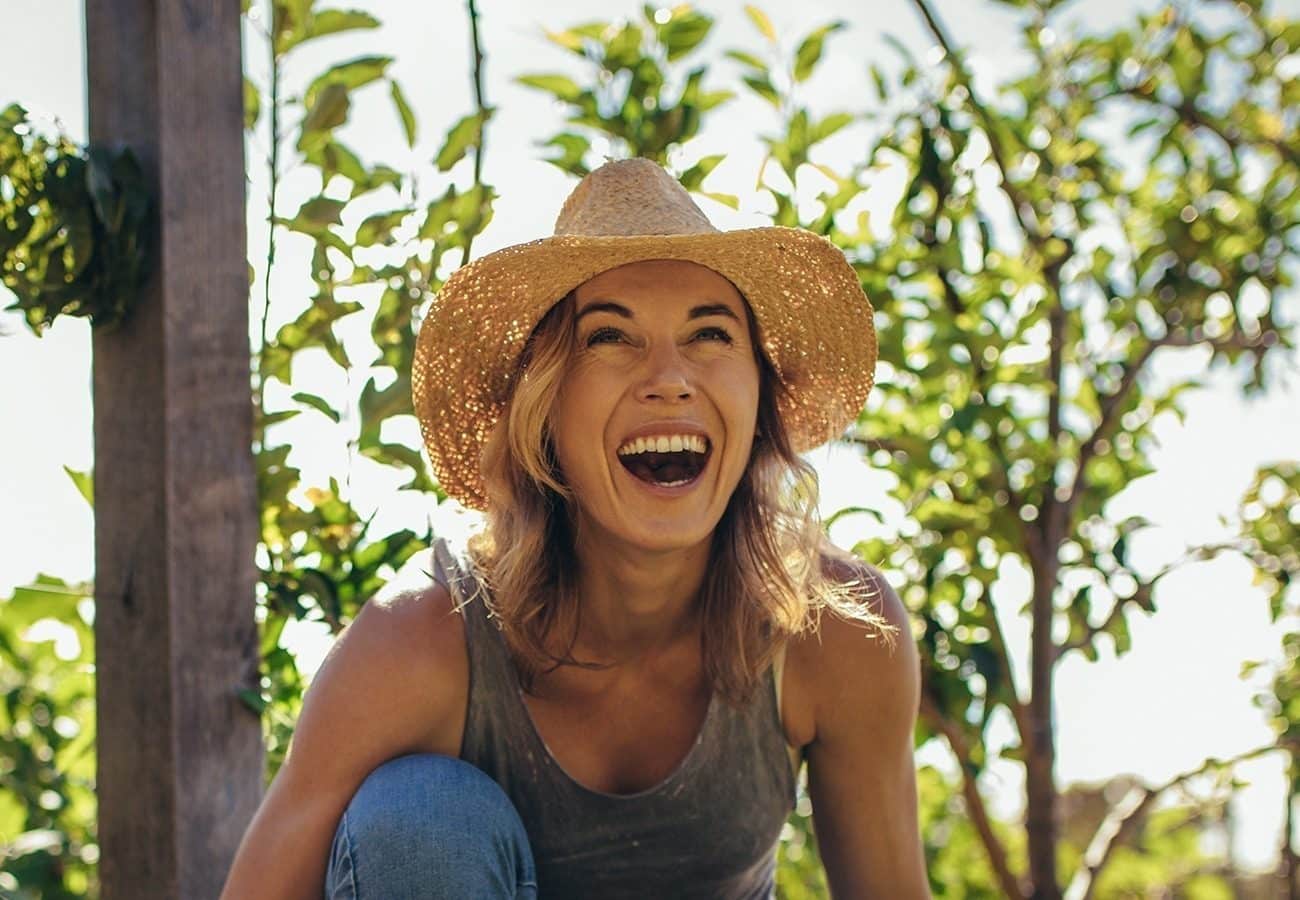 A woman wearing a straw hat and tank top smiles and laughs outdoors, surrounded by green leafy plants and sunshine.