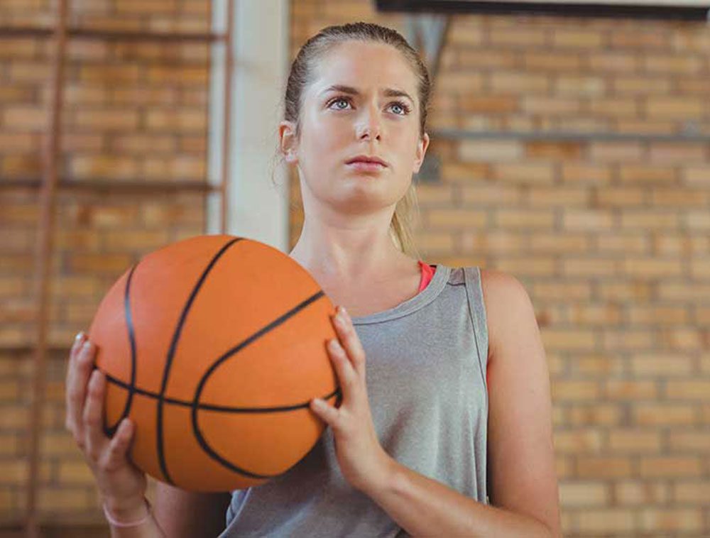 A young woman in a gray tank top holds a basketball with both hands, looking focused. She stands indoors in front of a blurred brick wall.