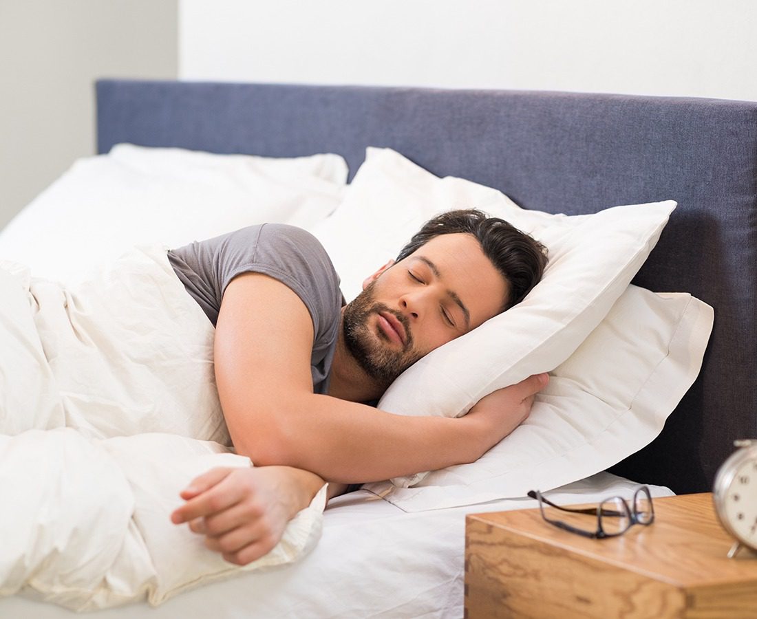 A man with dark hair and a beard is sleeping on his side in bed, covered by a white blanket. An alarm clock and eyeglasses are on the bedside table next to him.