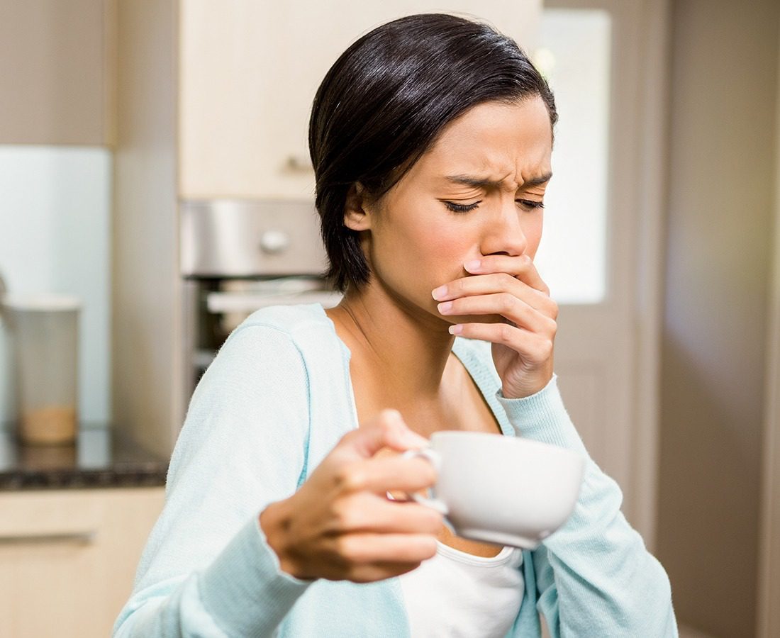 A woman in a light blue sweater holds a white cup in one hand and covers her mouth with the other, appearing to react with discomfort or nausea in a kitchen setting.