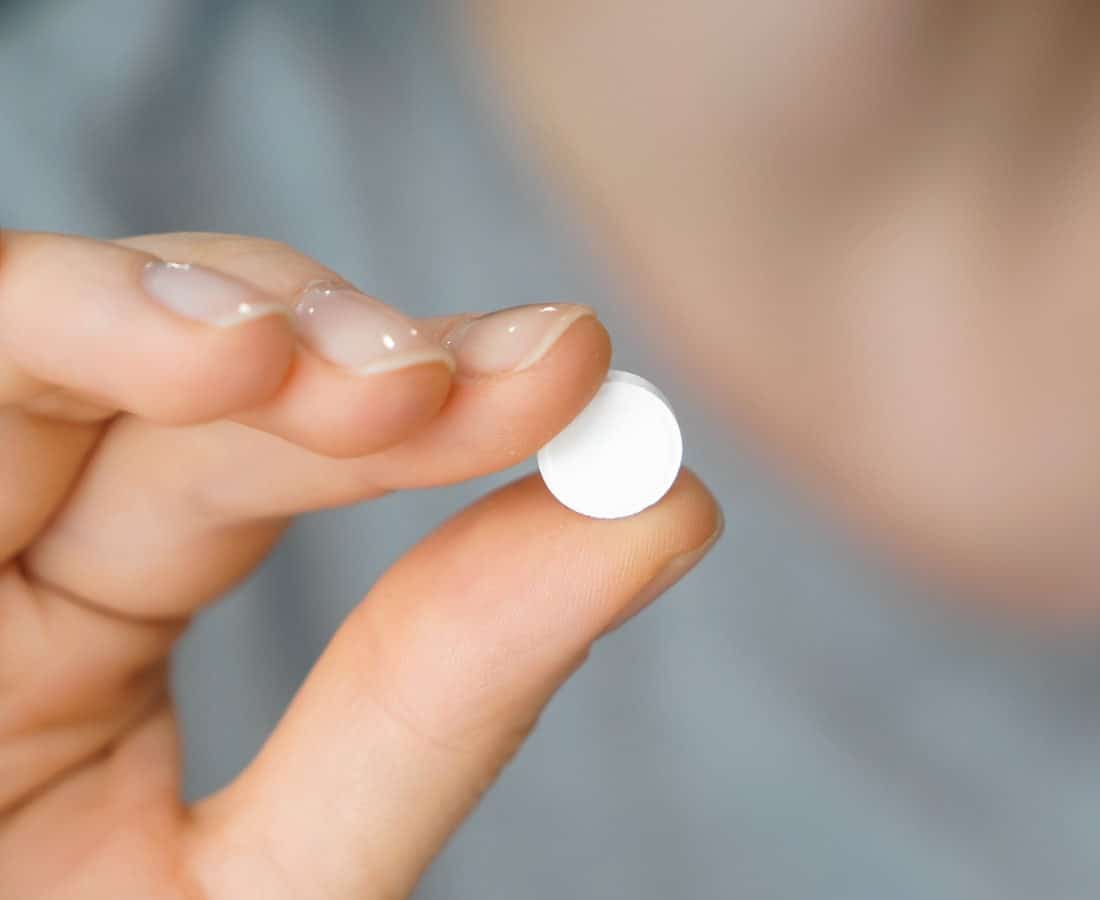 A close-up of a hand holding a single round white tablet between the thumb and index finger, with a blurred background.
