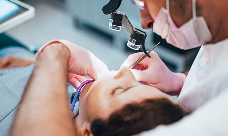 A dentist wearing magnifying glasses and gloves examines a patients mouth with dental tools. The patient is lying back in the dental chair during the procedure.