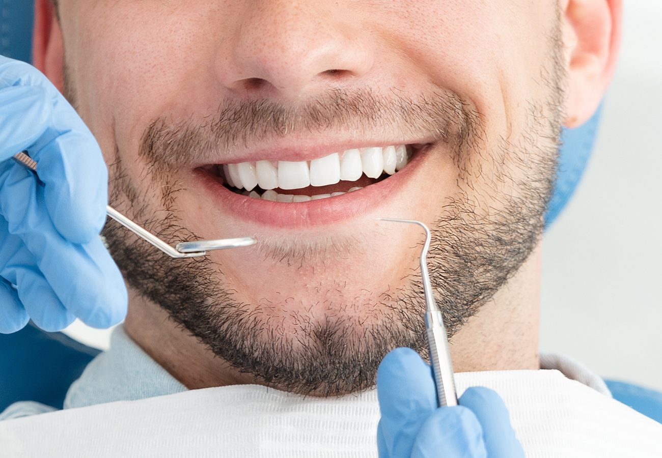 A close-up of a smiling man at the dentist, showing white teeth. Gloved hands hold dental instruments near his mouth, and a dental bib covers his chest.