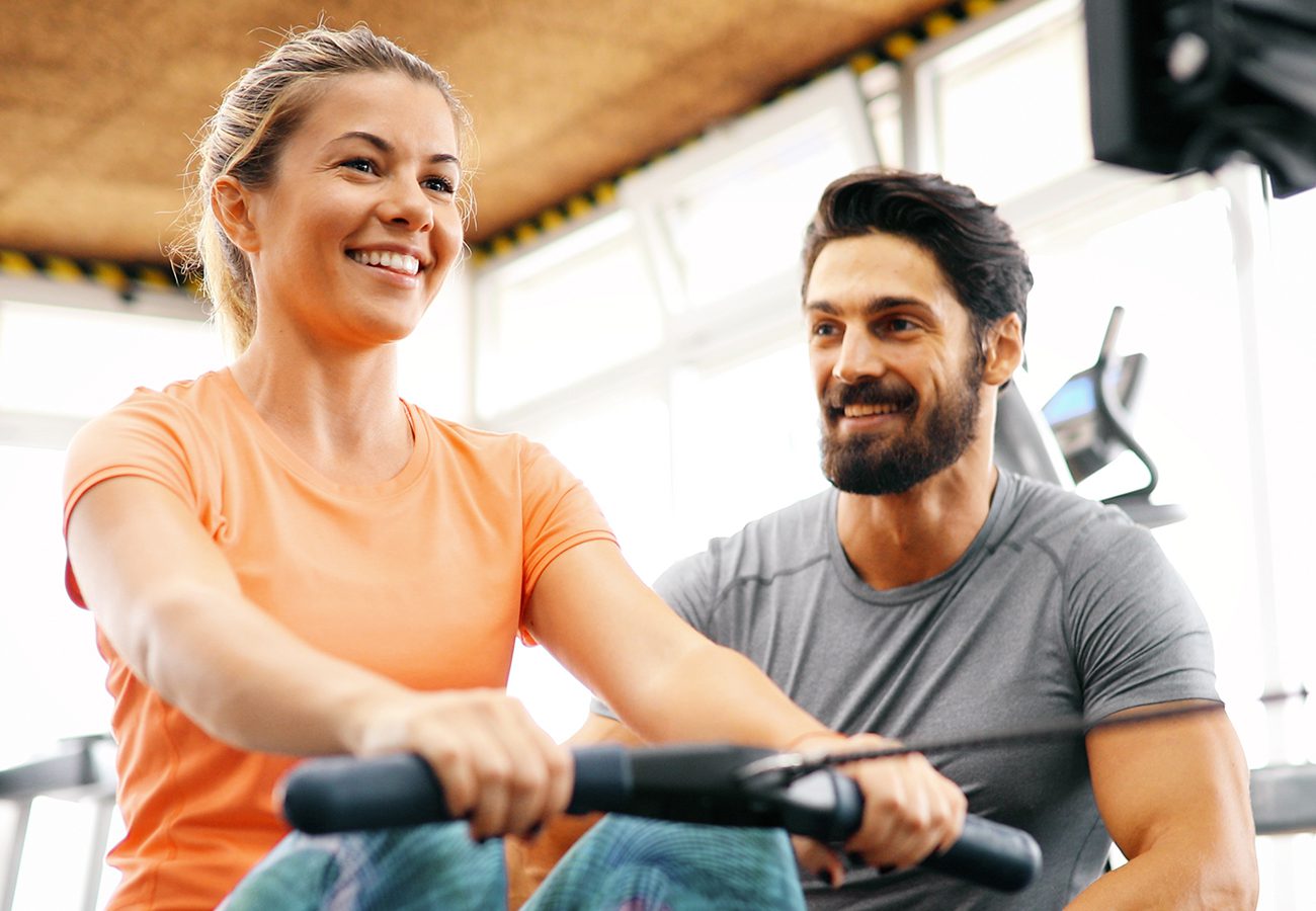 A woman in an orange shirt smiles while using a rowing machine at the gym; a man in a gray shirt sits beside her, watching and smiling supportively.