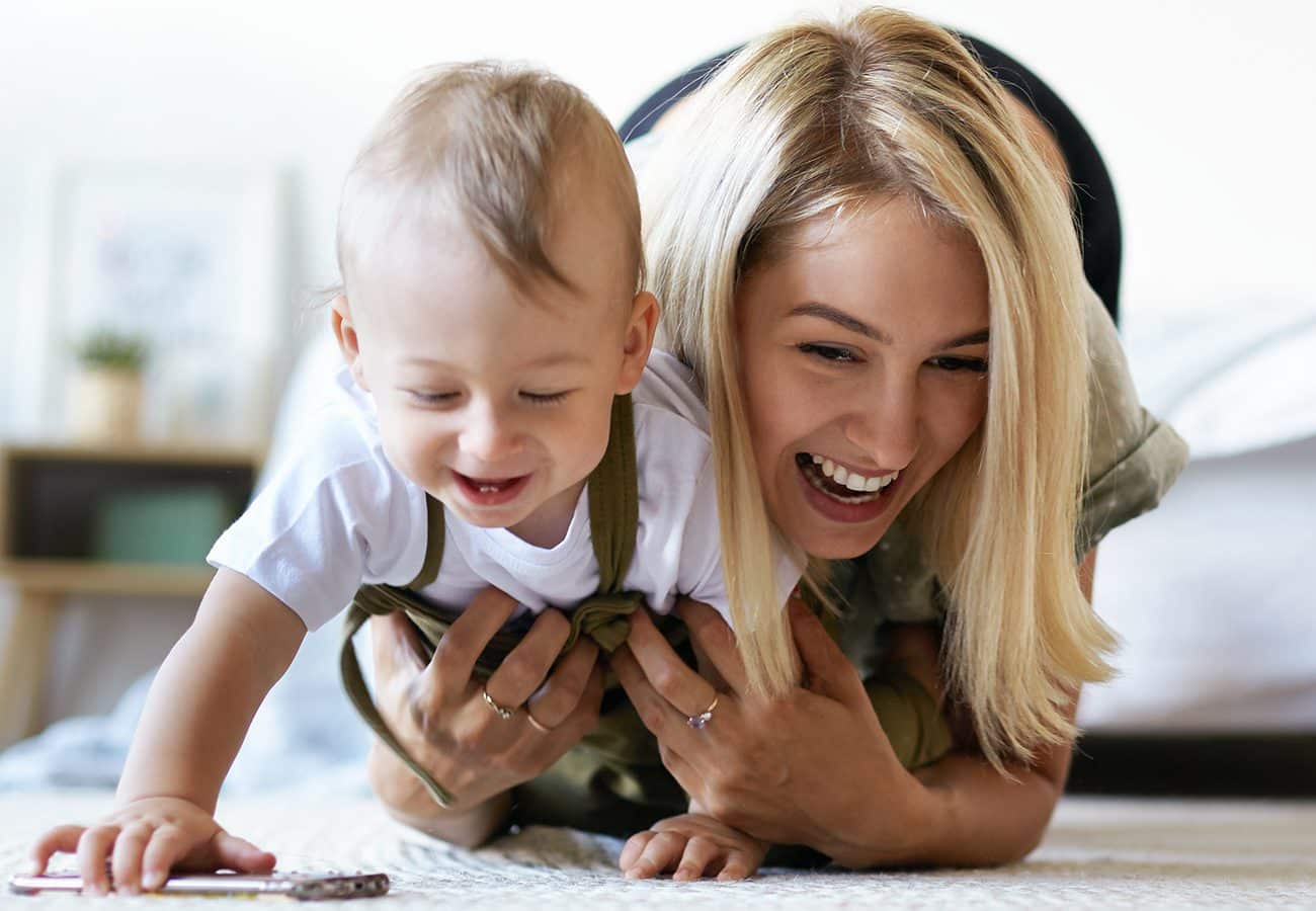 A smiling woman lies on the floor behind a laughing baby who is reaching forward. The woman appears to be helping the baby, and both look happy and playful in a bright, cozy indoor setting.