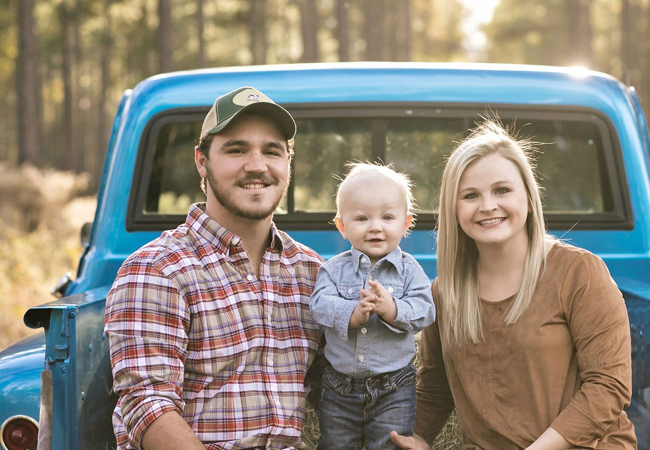 A man, woman, and small child pose smiling in front of an old blue pickup truck in a sunlit outdoor setting with trees in the background.
