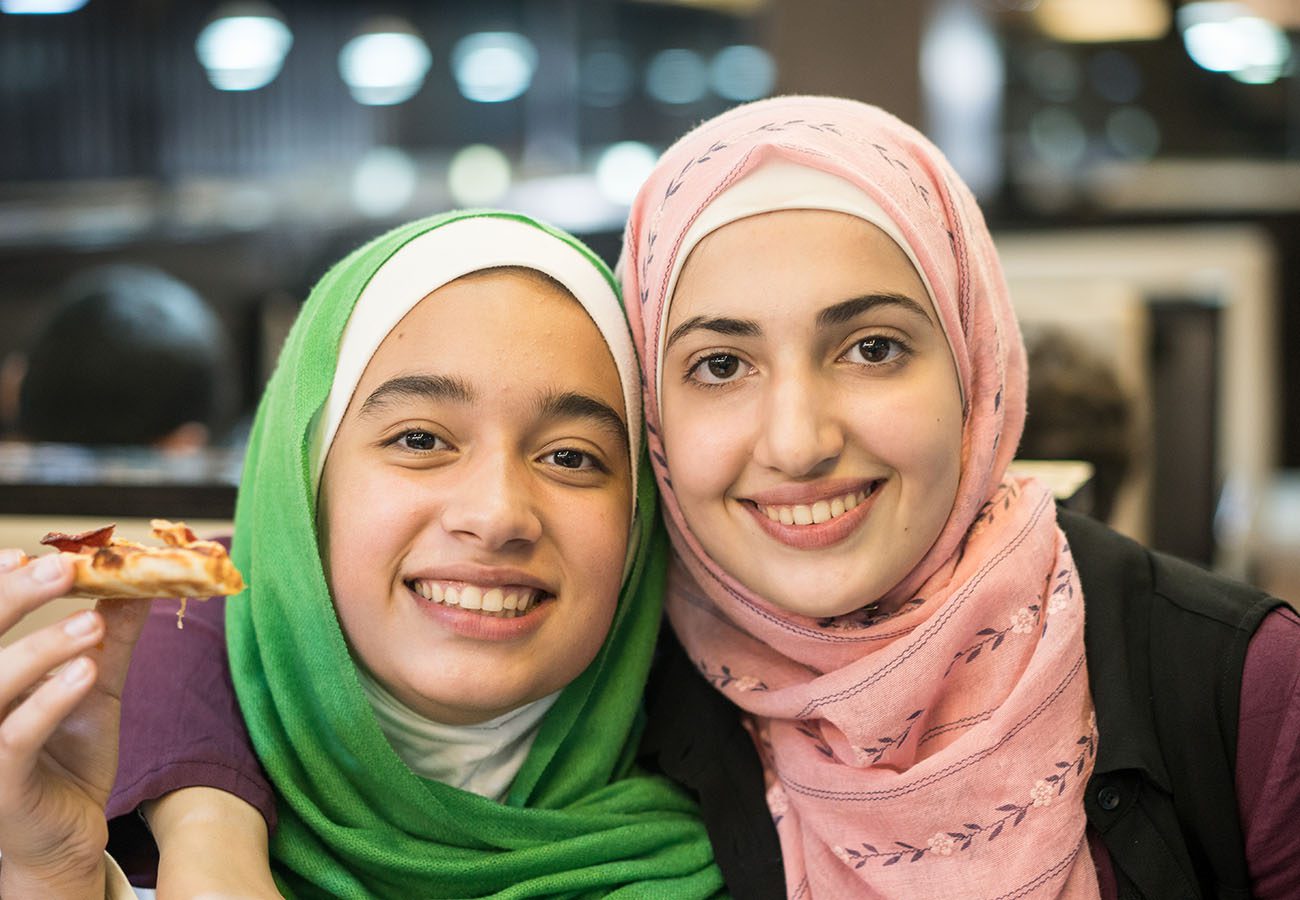 Two young women wearing hijabs, one green and one pink, smile closely together. One of them holds a slice of pizza. The background is softly blurred, suggesting they are in a restaurant.