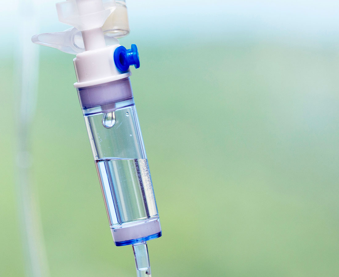 Close-up of a medical IV drip chamber partially filled with clear fluid, set against a blurred, light green background.
